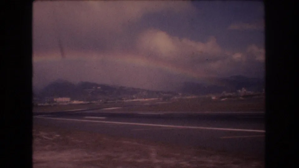 A 1977 Honolulu airport view from an aircraft window shows runway markings, distant hills, cloudy sky, and a faint rainbow over the landscape.