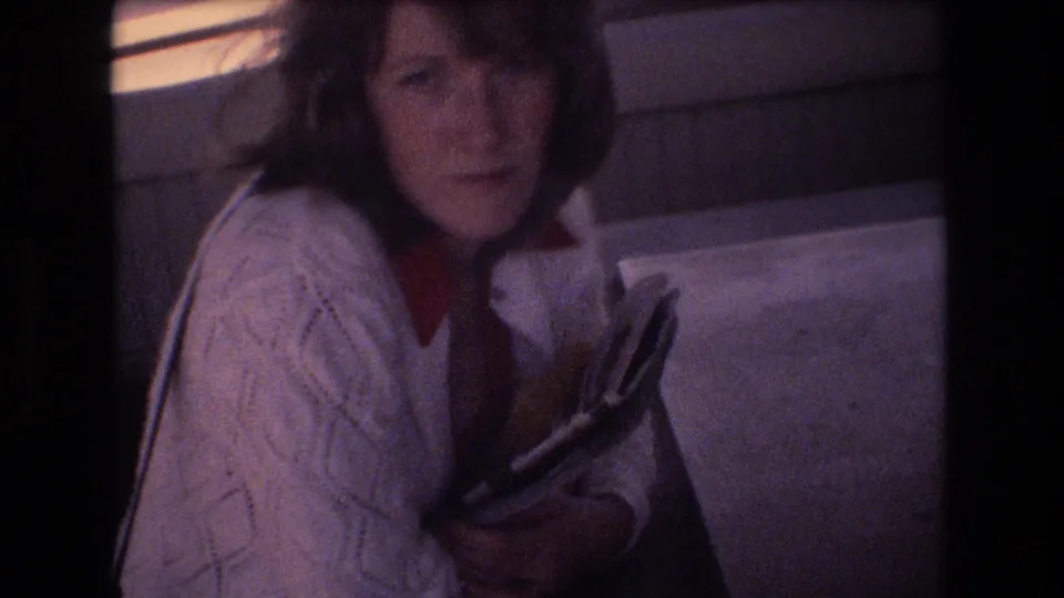Inside a 1977 airplane cabin, a woman in a white cable-knit sweater and red collar looks toward the camera while holding papers or a booklet.