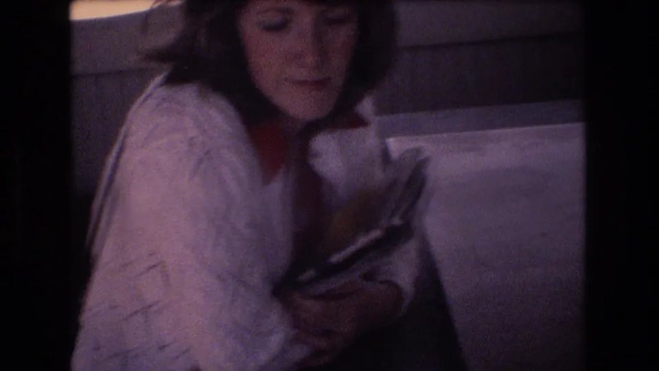 A woman seated in an airplane cabin in the 1977 Honolulu travel footage looks down at papers or a book, with the window area and seat back nearby.