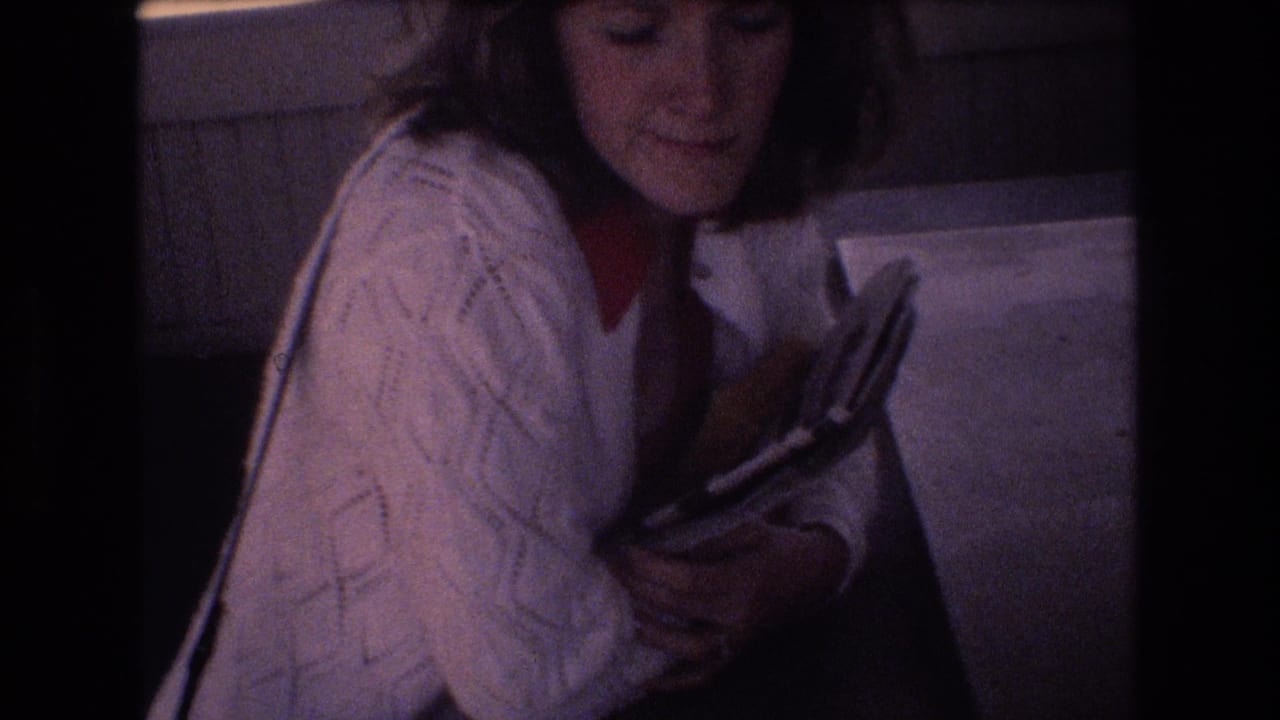 In 1977 Honolulu home-movie footage, a young woman with short dark hair wears a white cable-knit cardigan over a red blouse while holding a book or magazine indoors.