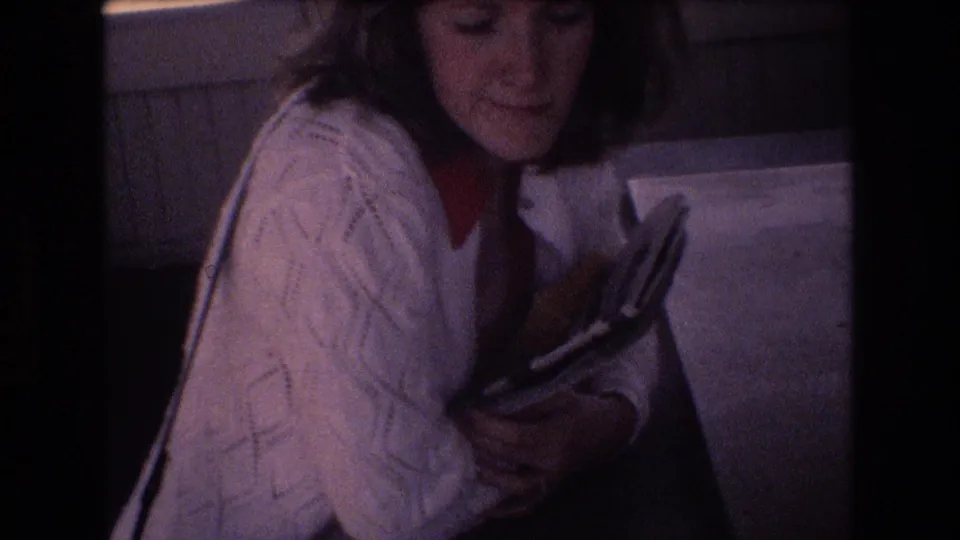 In 1977 Honolulu home-movie footage, a young woman with short dark hair wears a white cable-knit cardigan over a red blouse while holding a book or magazine indoors.