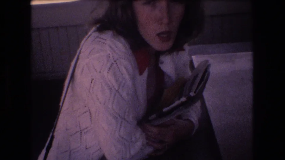 In the 1977 Honolulu interior scene, the woman appears to react or speak while clutching a book or magazine against her white cardigan.