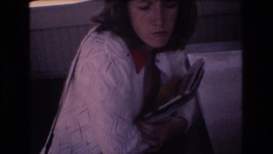 The 8mm frame shows the young woman seated indoors, looking down with a subdued expression as she holds a book or magazine close to her chest.