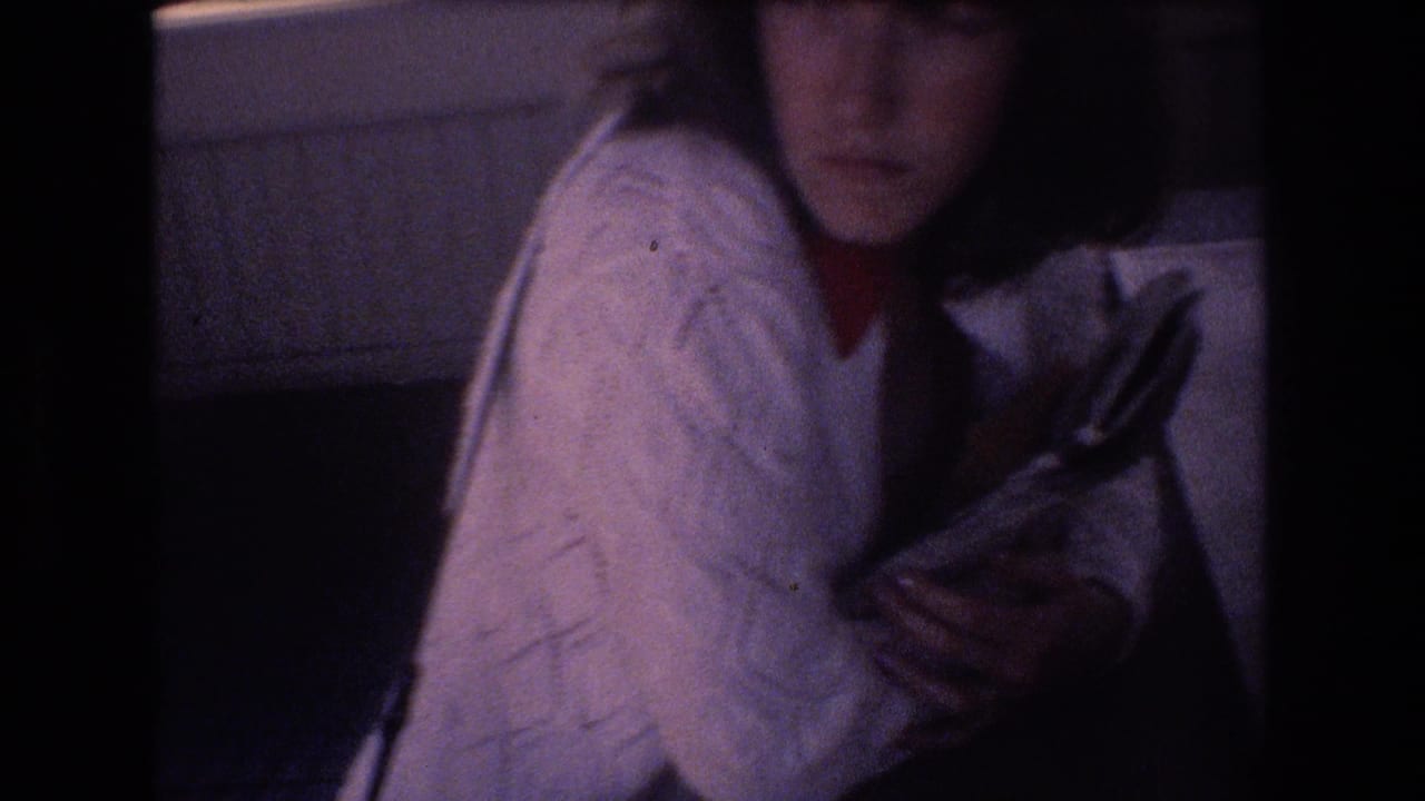 Young woman in a white cable-knit sweater and red collar holds books or magazines in a dim 1977 Honolulu home-movie interior.
