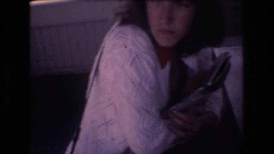 Young woman in a white cable-knit sweater and red collar holds books or magazines in a dim 1977 Honolulu home-movie interior.