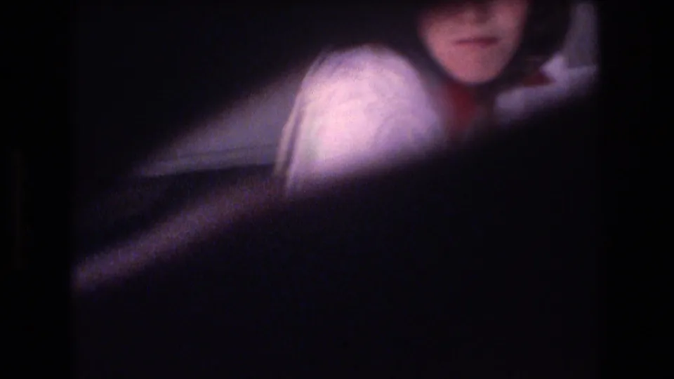 A woman in a light sweater is partially visible behind a dark foreground obstruction in soft, underexposed 8mm footage.