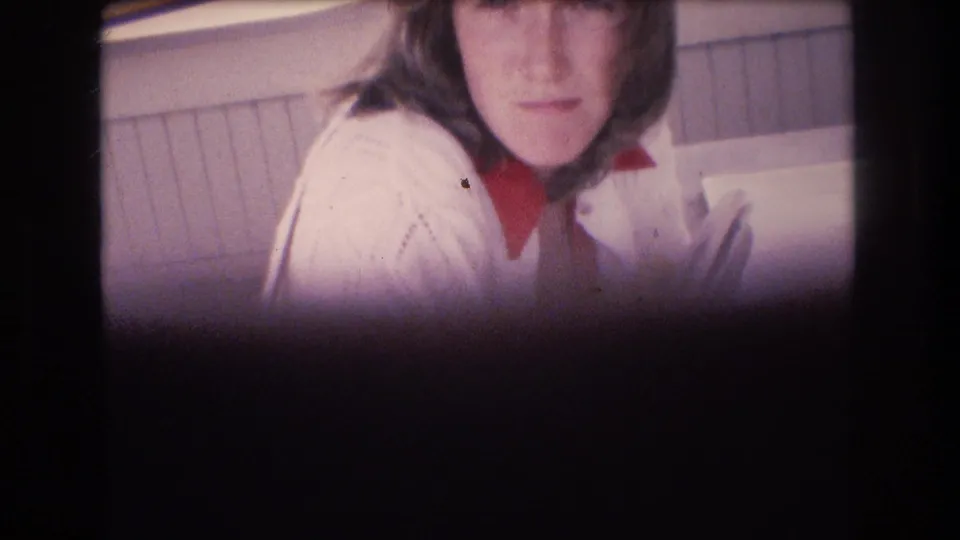 Closer home-movie frame of the young woman in a white sweater and red turtleneck holding books or magazines near a paneled wall.