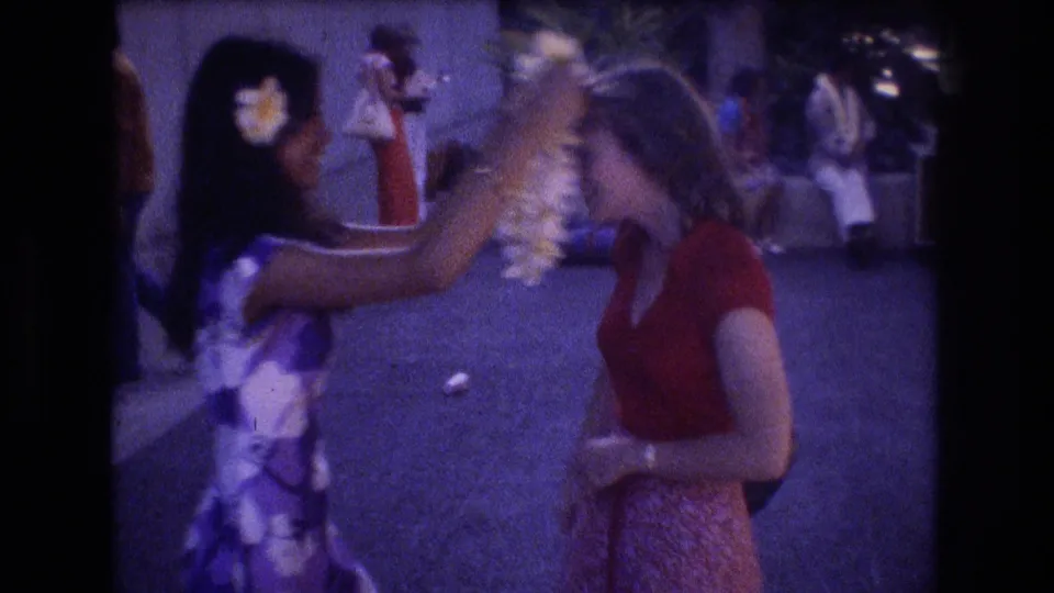 In the metadata-supported Honolulu 1977 scene, a woman in a purple and white dress places a flower lei over a woman wearing a red top.