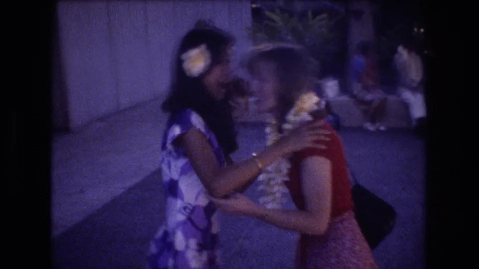 Two women with flower leis greet closely outdoors in the 1977 Honolulu home-movie clip, with other people and pavement in the background.
