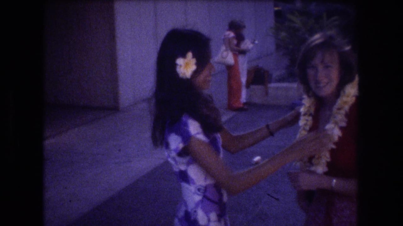In 1977 Honolulu home-movie footage, a young woman in a purple floral dress and plumeria hair flower greets another woman wearing red by placing or adjusting a white lei.