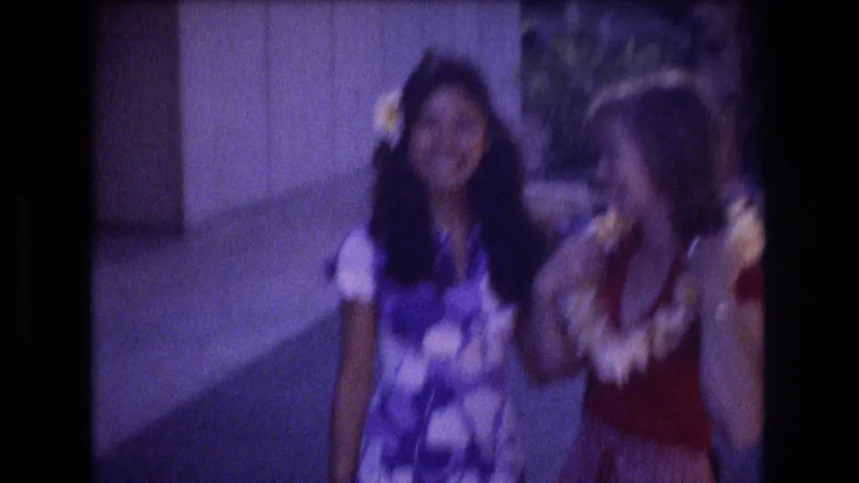 Two young women in a 1977 Honolulu home-movie frame smile and lean together, with one wearing a white lei and the other wearing a plumeria flower in her hair.