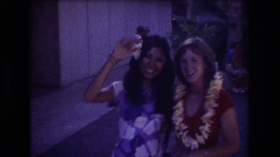 In the Honolulu 1977 home-movie shot, the two young women pose close together with a lei visible as one raises a hand in a cheerful wave.