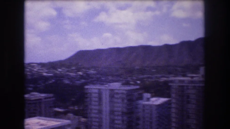 1977 8mm elevated Honolulu city view showing high-rise hotel or condominium buildings, cloudy sky, and Diamond Head in the distance.