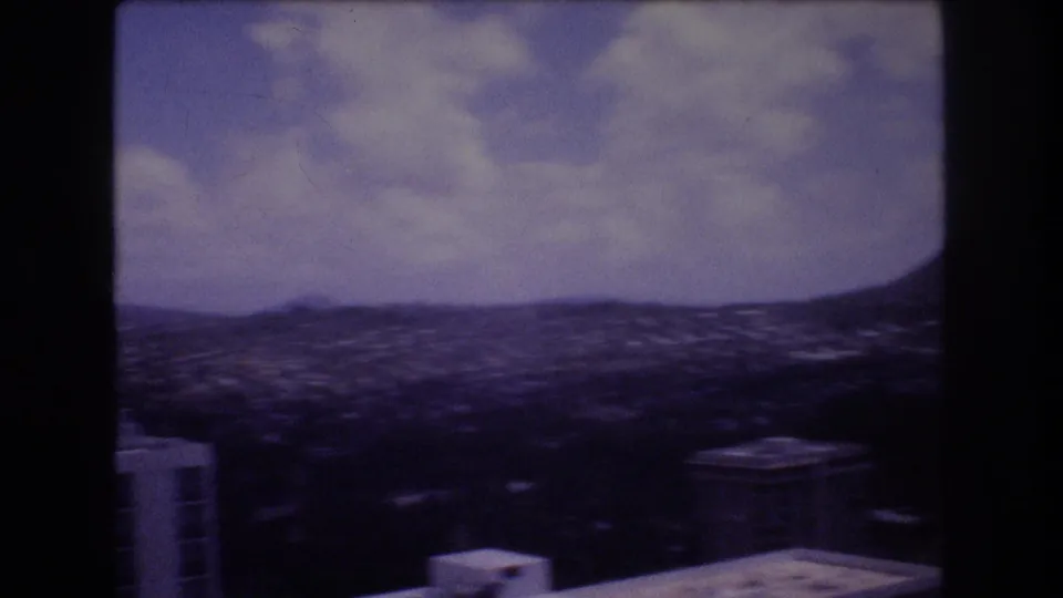 Wide 1977 Honolulu panorama with low-rise city areas, distant mountain ridges, cloudy sky, and cropped building rooftops in the foreground.