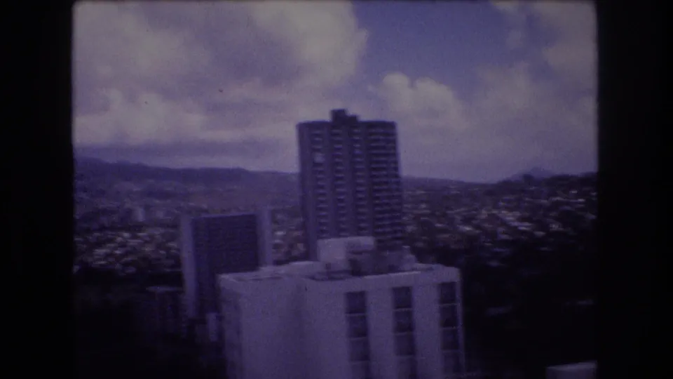 1977 8mm Honolulu view with a tall high-rise tower, foreground hotel or condominium structures, dense city blocks, distant hills, and cloudy sky.