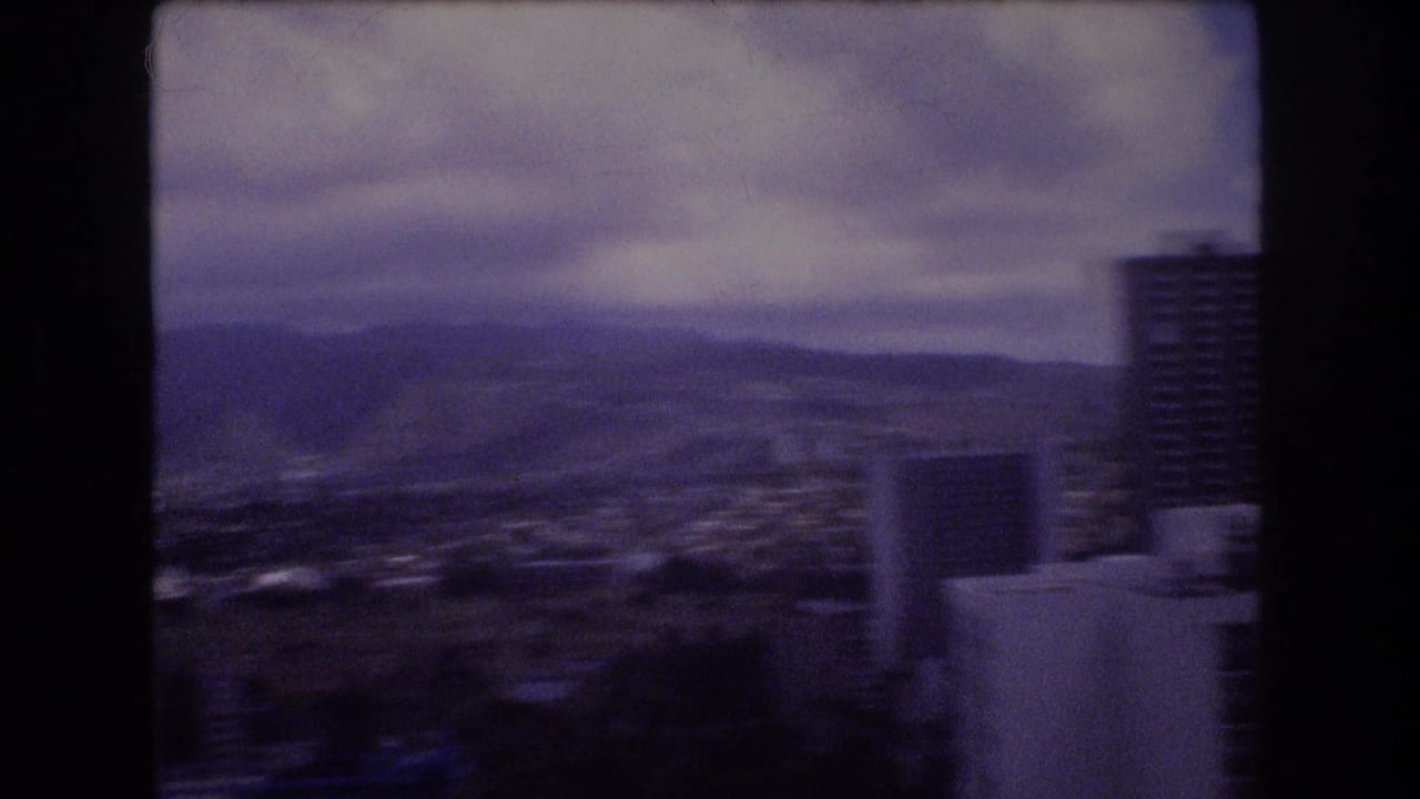 1977 8mm Honolulu home-movie frame showing a cloudy elevated view across city buildings toward distant mountain ridges, with a dark vertical frame edge at right.