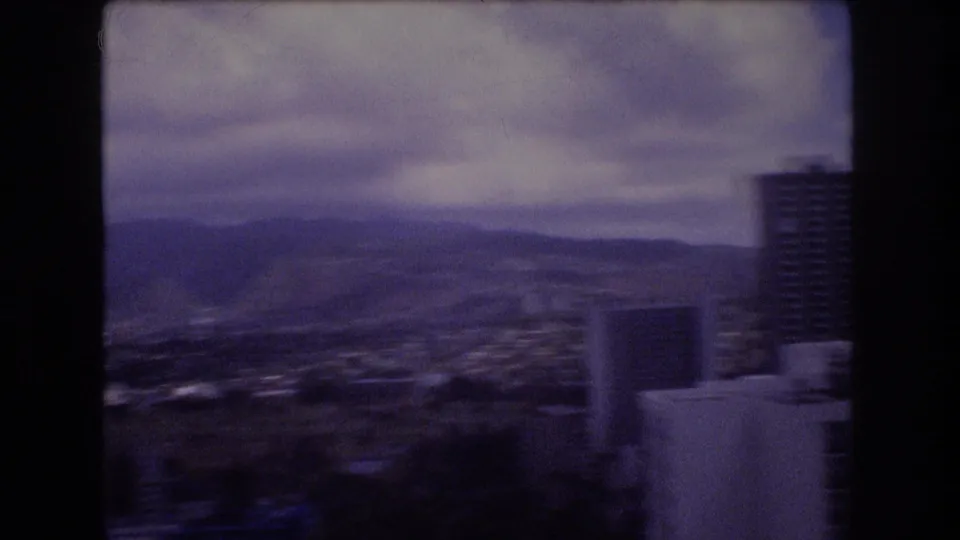 1977 8mm Honolulu home-movie frame showing a cloudy elevated view across city buildings toward distant mountain ridges, with a dark vertical frame edge at right.