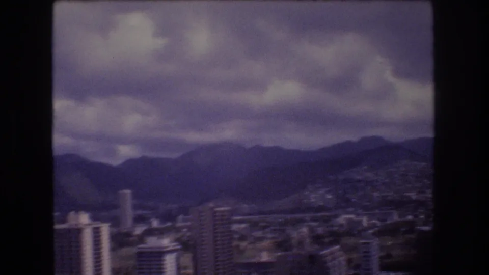 1977 8mm Honolulu frame with several towers in the foreground, residential areas beyond, and dark mountain slopes under layered clouds.