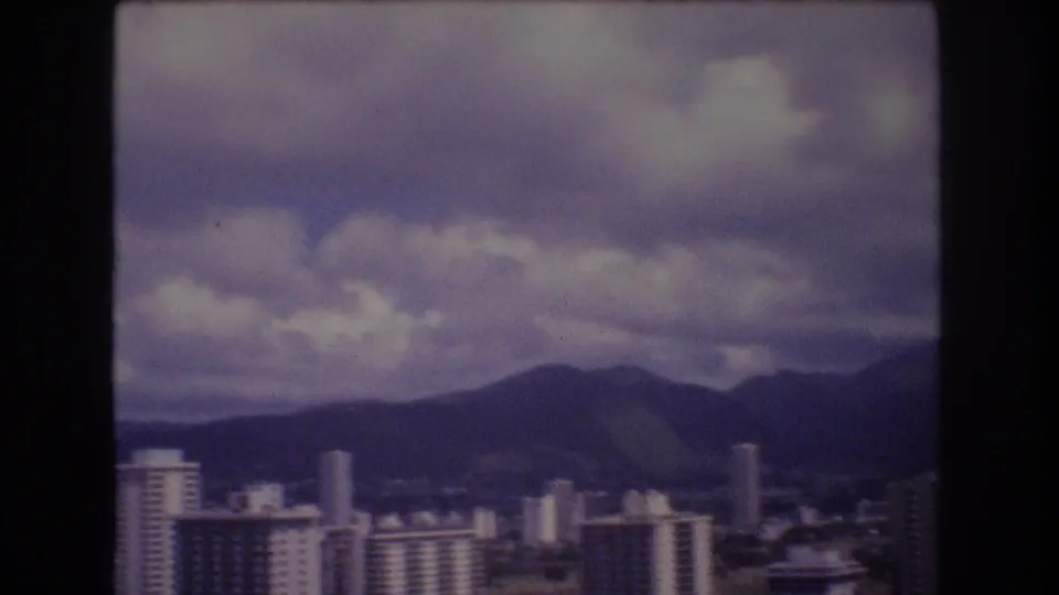 1977 8mm Honolulu skyline view showing pale high-rise buildings and a rounded mountain ridge under a textured cloudy sky.