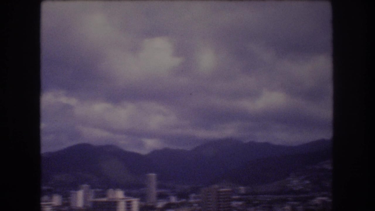 1977 Honolulu skyline frame showing low and mid-rise city buildings beneath a dark mountain ridge and layered overcast clouds.