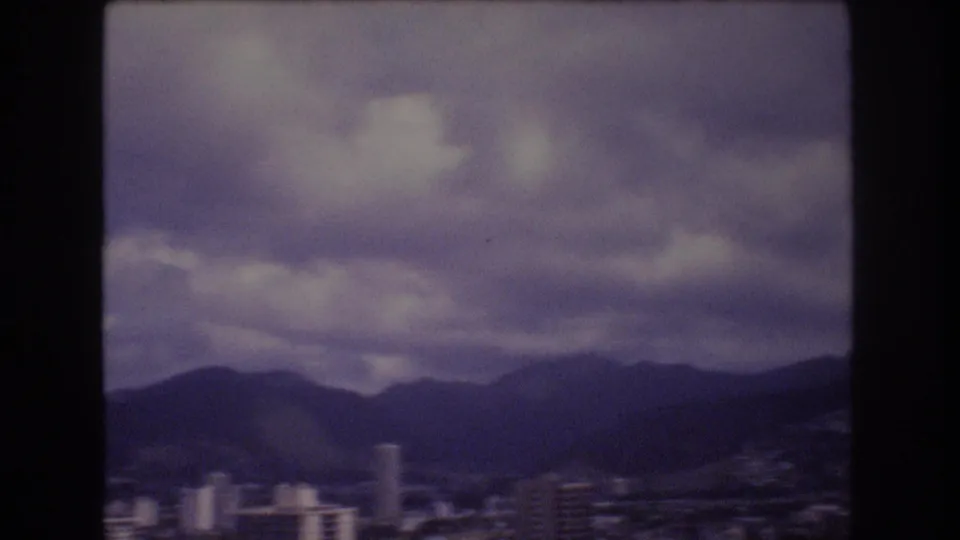 1977 Honolulu skyline frame showing low and mid-rise city buildings beneath a dark mountain ridge and layered overcast clouds.