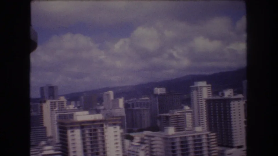 1977 Honolulu skyline frame with multiple city buildings, a sloping hill behind them, and bright cloud cover across the upper frame.