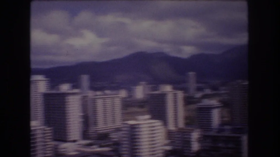 1977 Honolulu cityscape frame showing a slightly blurred cluster of high-rises in front of dark hills and overcast sky.