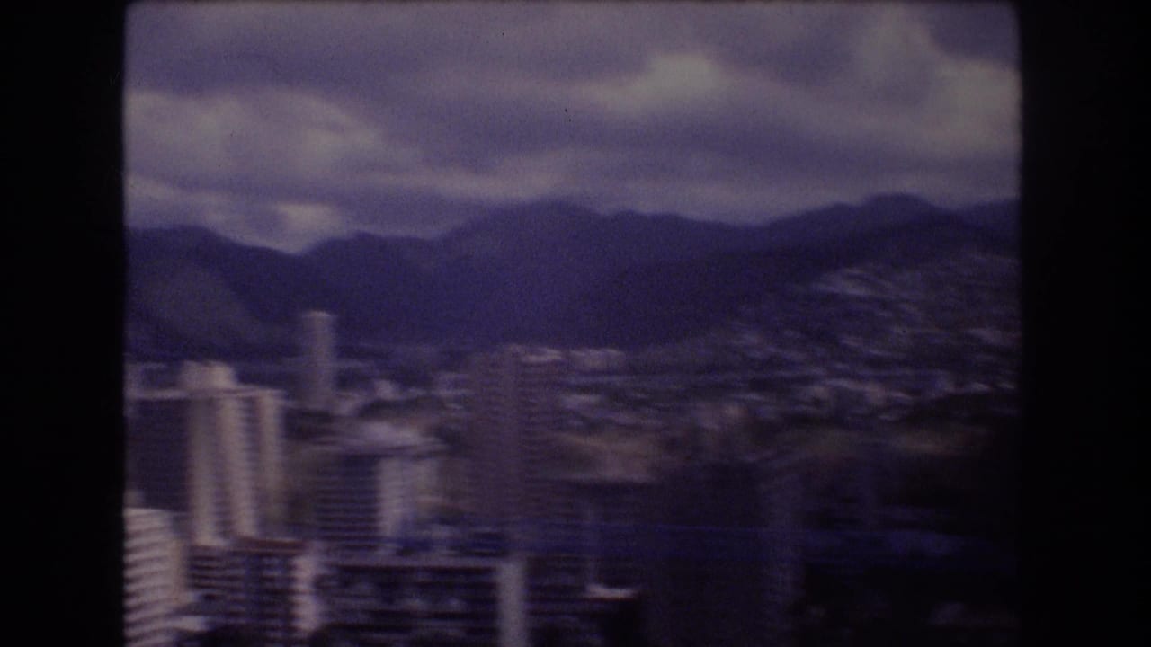 1977 8mm frame of Honolulu, Hawaii, with high-rise buildings and a broad city area below dark mountains under cloud cover.