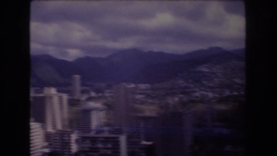 1977 8mm frame of Honolulu, Hawaii, with high-rise buildings and a broad city area below dark mountains under cloud cover.