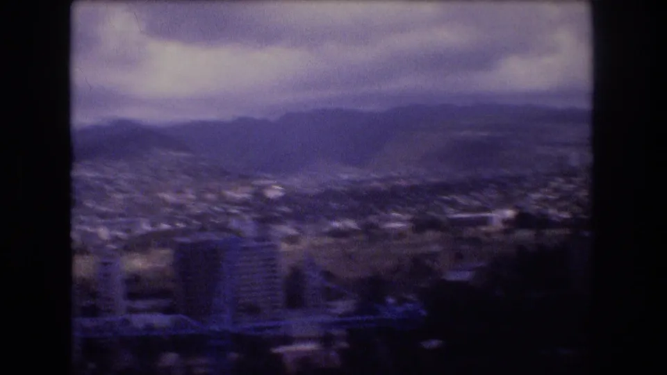 1977 8mm Honolulu frame with a broad cityscape, distant mountain ridge, low clouds, and soft motion blur from the moving camera.