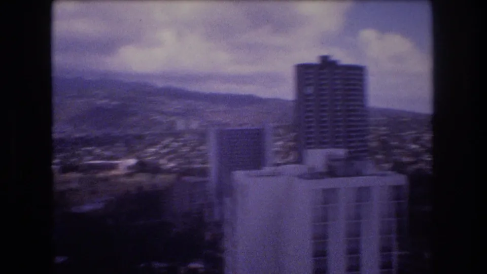 1977 Honolulu frame showing a prominent high-rise in the foreground, lower urban buildings, distant terrain, and a cloudy sky.