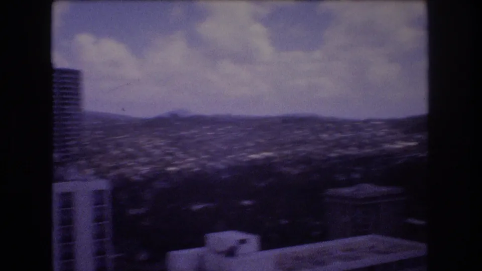 1977 Honolulu city view with partial high-rise structures at the edges, a broad urban valley, distant hills, and visible 8mm gate borders.