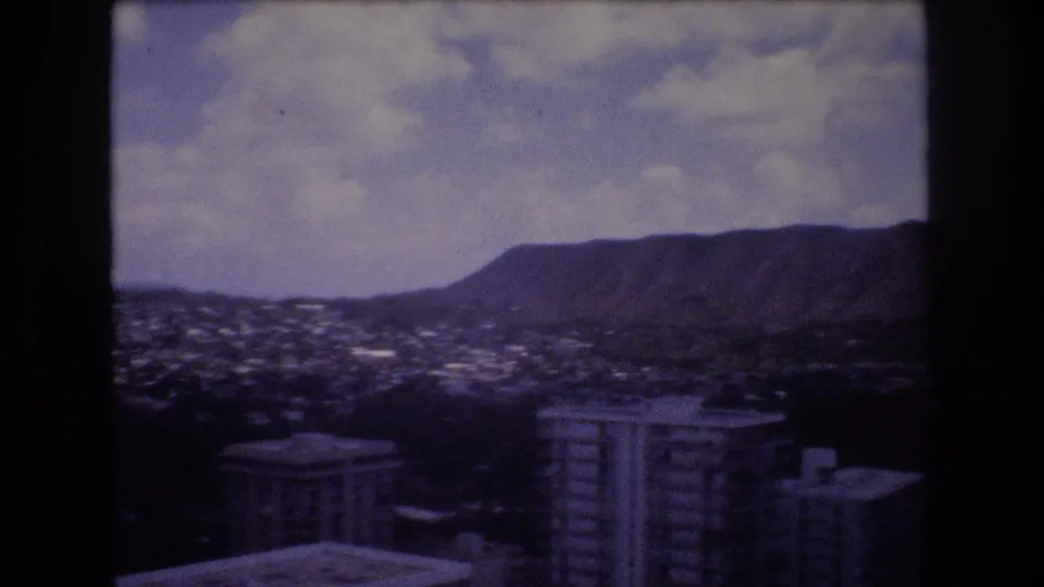 1977 8mm frame showing Honolulu's urban valley, foreground apartment or hotel towers, and a long mountain ridge under broken cloud cover.
