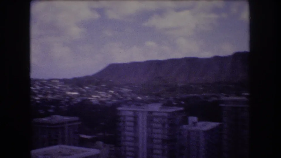 Late-1970s Honolulu home-movie frame with high-rise rooftops, a sprawling city valley, and a prominent mountain ridge in soft purple-toned 8mm color.