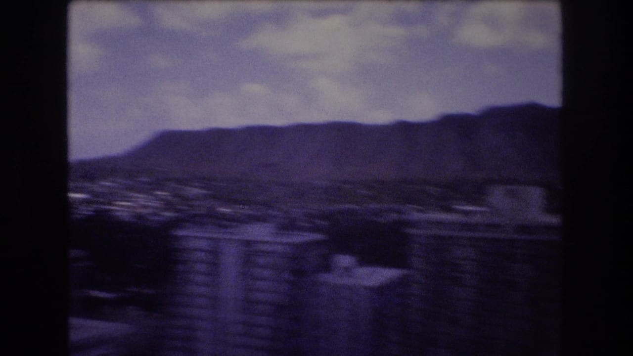 1977 Honolulu home-movie frame showing marina water in the foreground and the Diamond Head ridgeline in the distance under a cloudy sky.