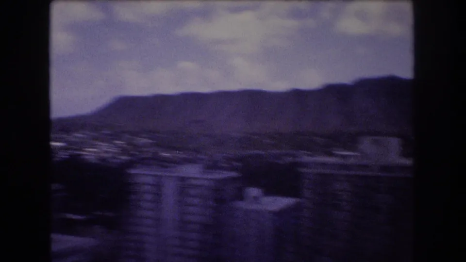 1977 Honolulu home-movie frame showing marina water in the foreground and the Diamond Head ridgeline in the distance under a cloudy sky.