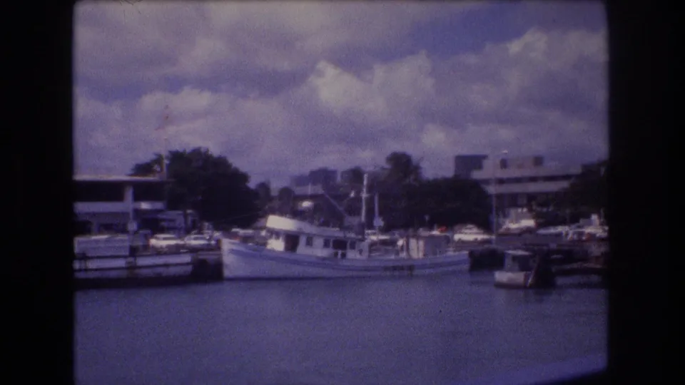 1977 Honolulu marina view with a white boat tied near docks, waterfront structures, parked cars, trees, and clouds above the harbor.