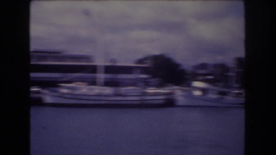 Motion-blurred 1977 Honolulu waterfront frame showing boats along the marina, water in the foreground, and low urban structures during a handheld pan.