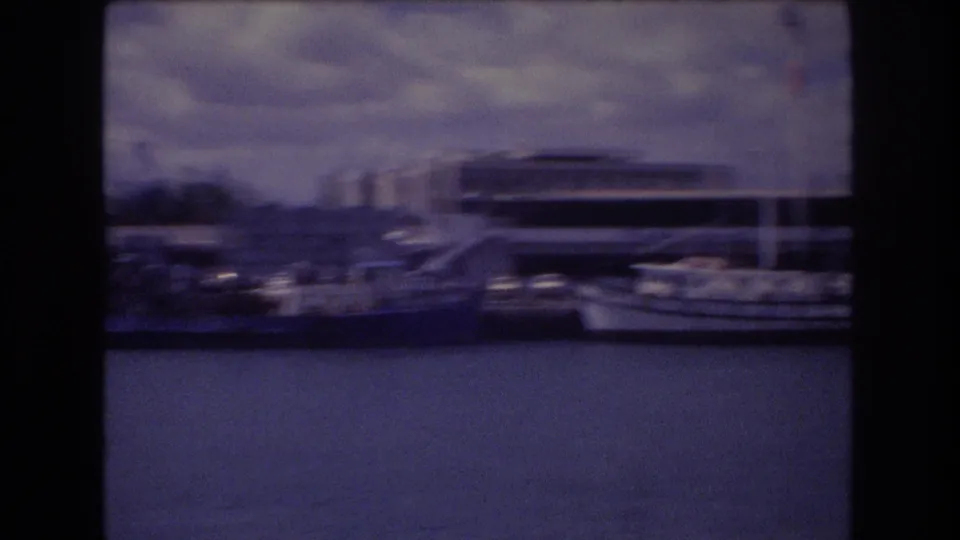 1977 Honolulu marina frame with several moored boats, calm harbor water, a modern waterfront building, distant high-rises, and cloudy sky.
