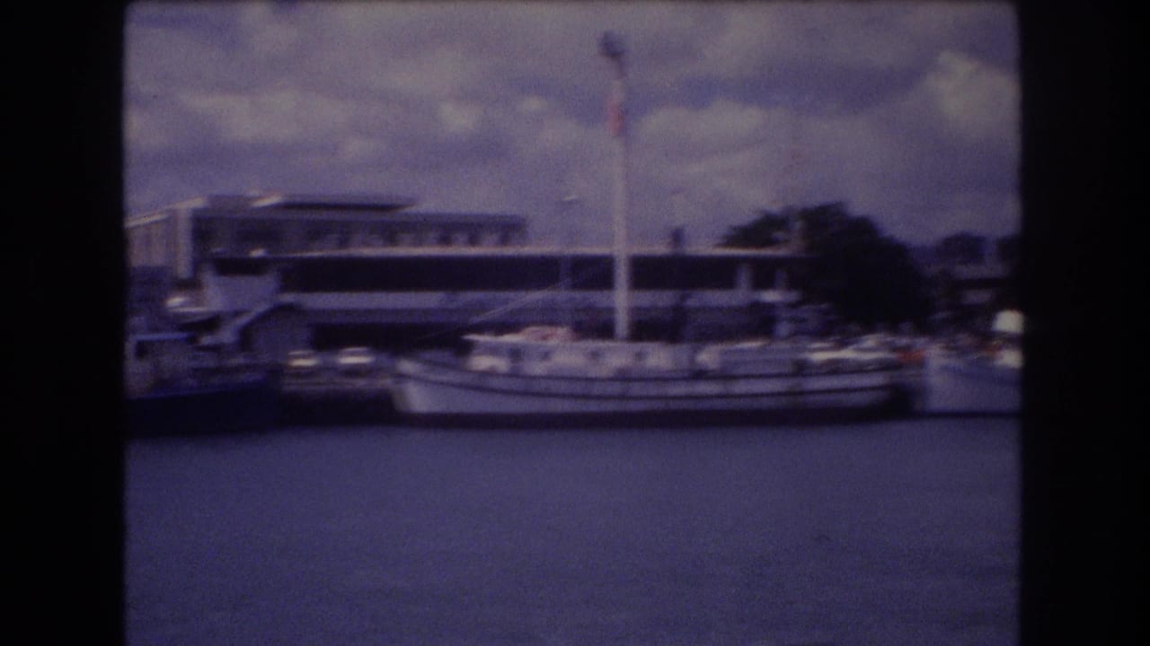 1977 Honolulu harbor view from a boat, showing docked vessels, a larger white ferry-like craft, waterfront buildings, and a cloudy sky.