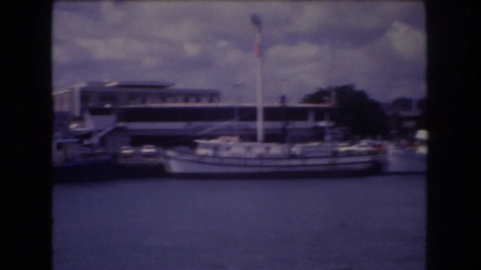 1977 Honolulu harbor view from a boat, showing docked vessels, a larger white ferry-like craft, waterfront buildings, and a cloudy sky.