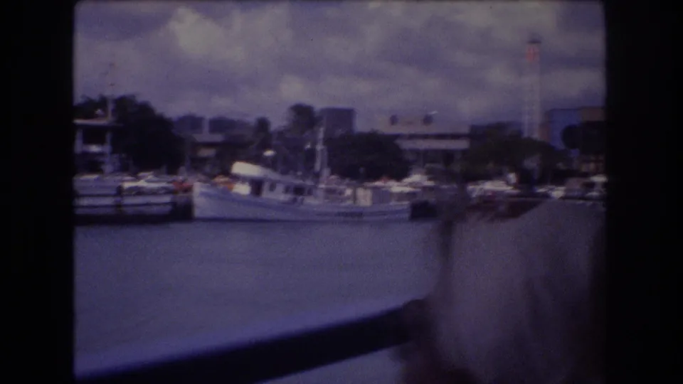 1977 Honolulu boat-tour view across harbor water, with vessels moored near waterfront buildings and a passenger partly visible at the right edge.