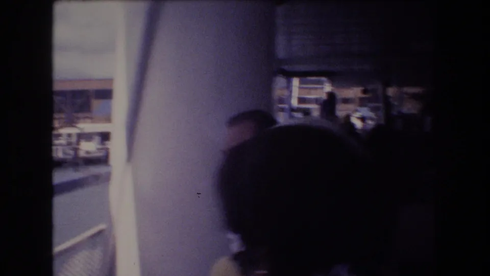 Interior or covered area of a 1977 Honolulu tour boat, with passengers in shadow, a column, a window opening, and harbor scenery beyond.