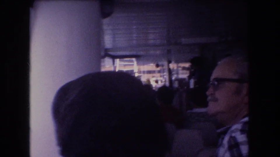 Adult passengers on a 1977 Honolulu boat tour, including a man wearing glasses, seated or standing near a column while looking toward the harbor.