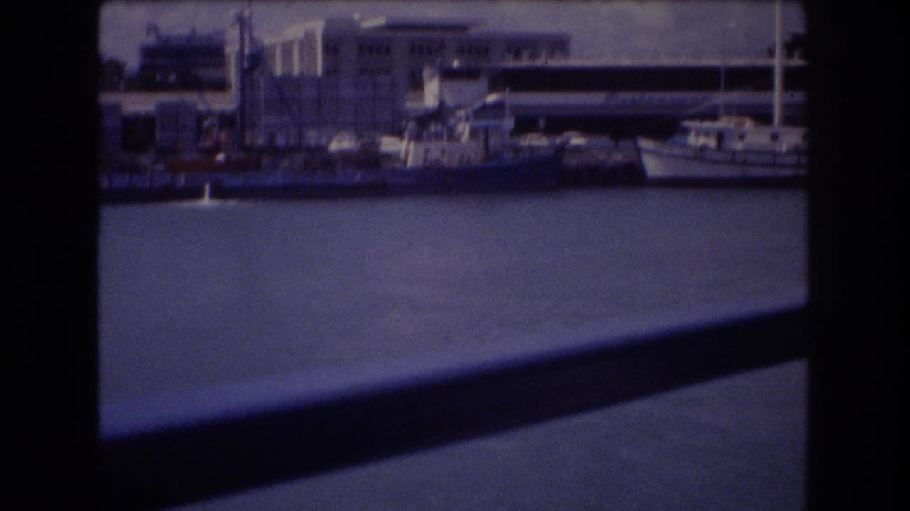 1977 Honolulu harbor shoreline appears in soft 8mm color, with boats, water, waterfront buildings, and dark foreground bars from a shipboard viewpoint.