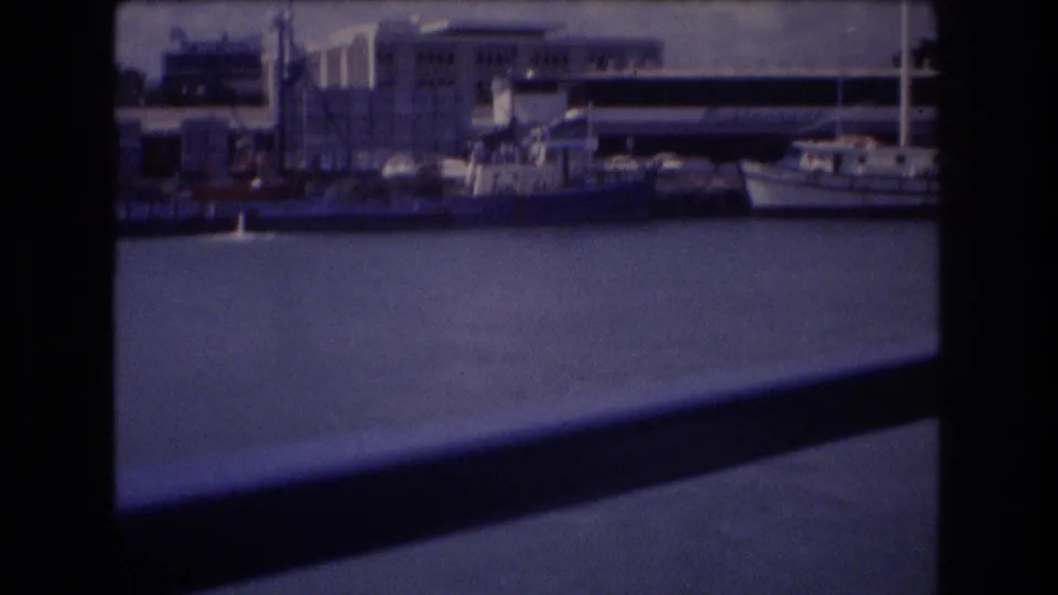 1977 Honolulu harbor shoreline appears in soft 8mm color, with boats, water, waterfront buildings, and dark foreground bars from a shipboard viewpoint.