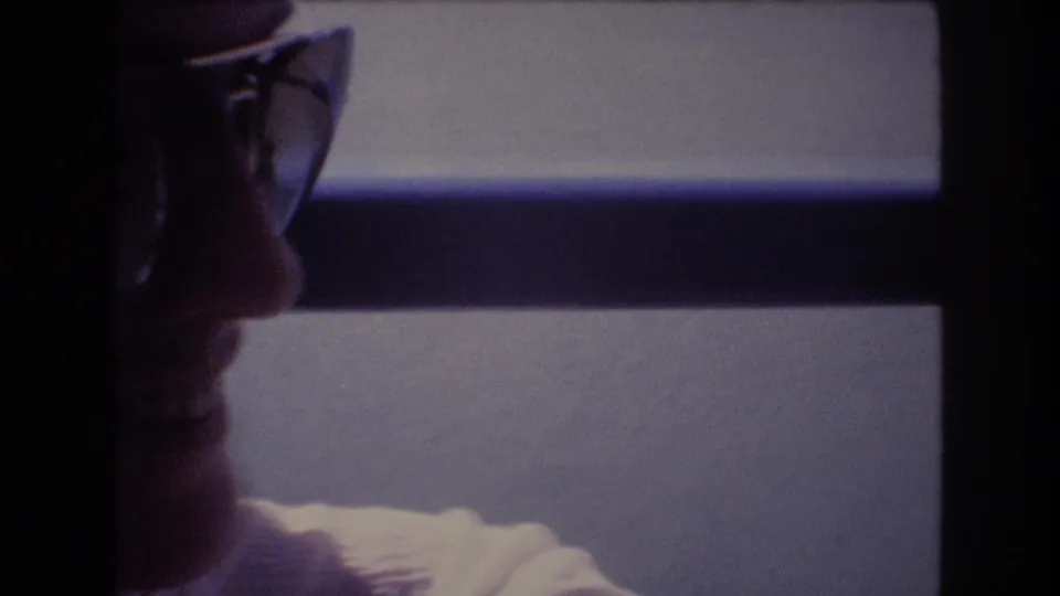 A man wearing glasses is shown in profile inside the vessel, looking toward the water through a dark-framed window in grainy 8mm footage.