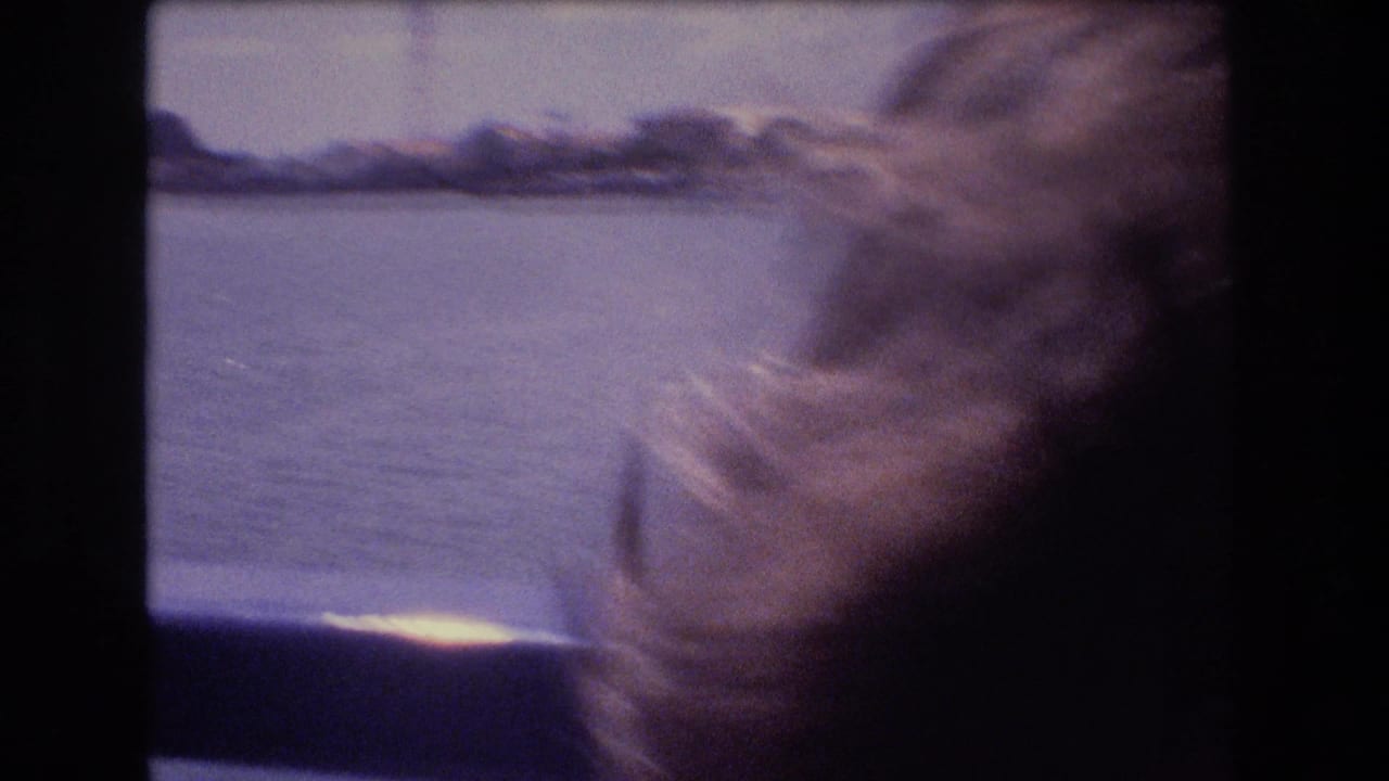Windblown woman near a boat window with water and shoreline outside.