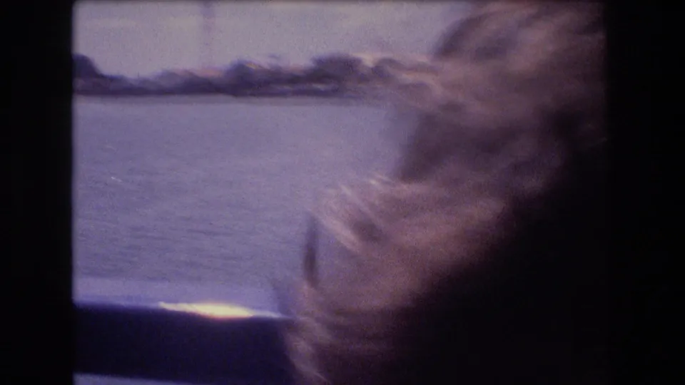 Windblown light hair fills the right side of the frame as water, distant shoreline, and a boat-window frame appear outside in the Honolulu travel footage.