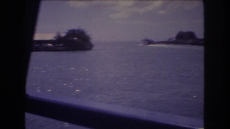 A boat-window view looks across rippled water toward a small palm-lined shoreline and low landforms in 1977 Honolulu home-movie footage.